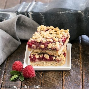 Two raspberry bars stacked on a square white plate with fresh raspberries and mint to garnish