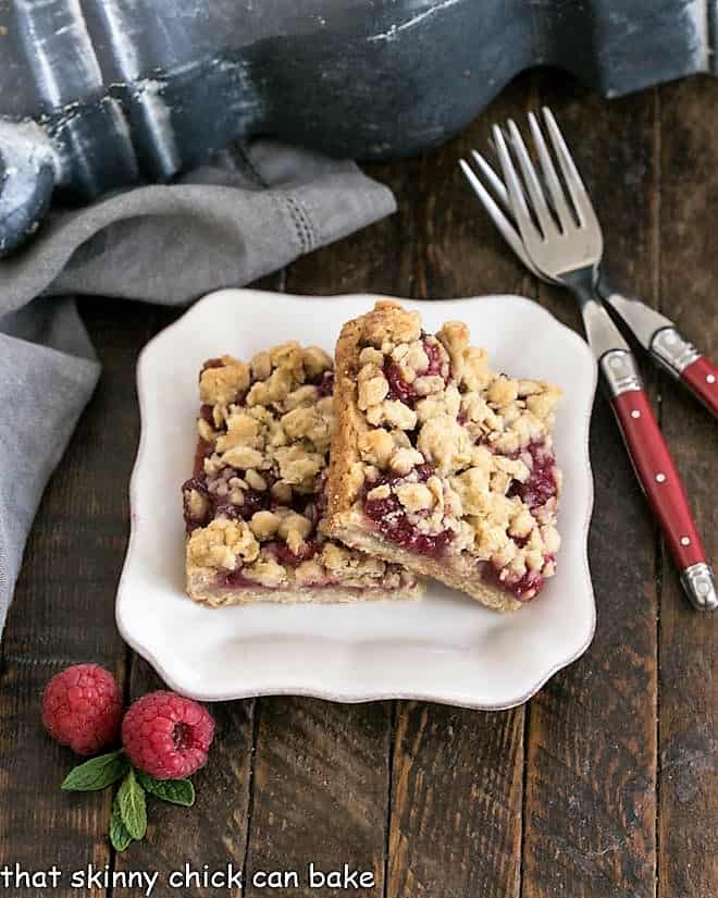 Two rasbperry Crumble bars on a white dessert plate with 2 red handled forks.