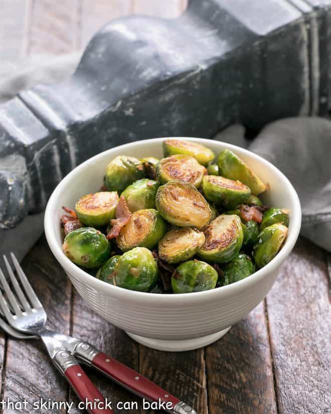A white bowl of skillet brussels sprouts with bacon and two red handled forks