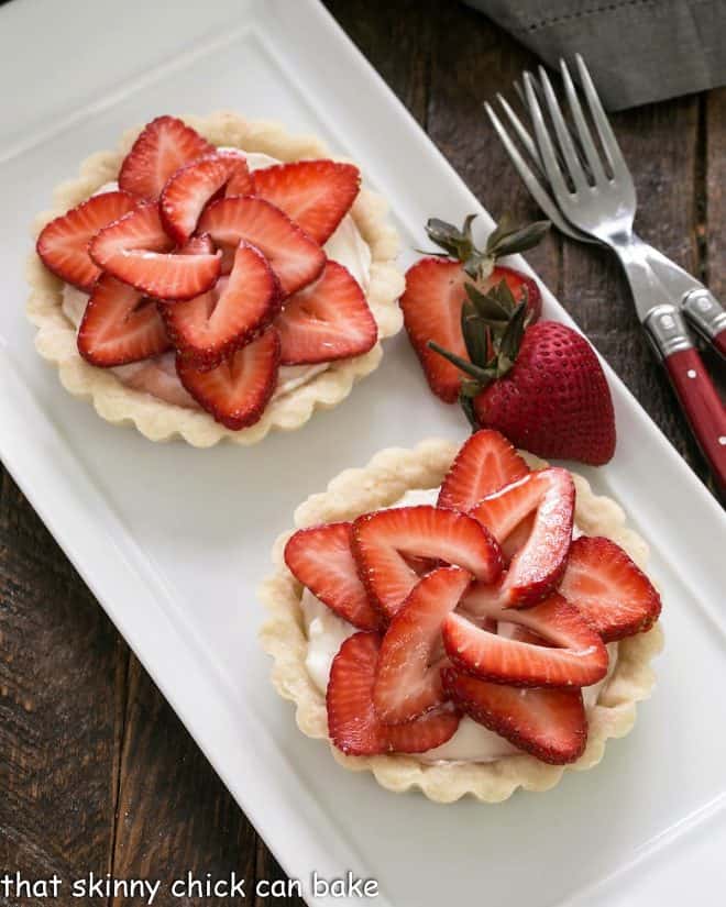 2 fresh strawberry tarts on a white ceramic tray with a strawberry garnish