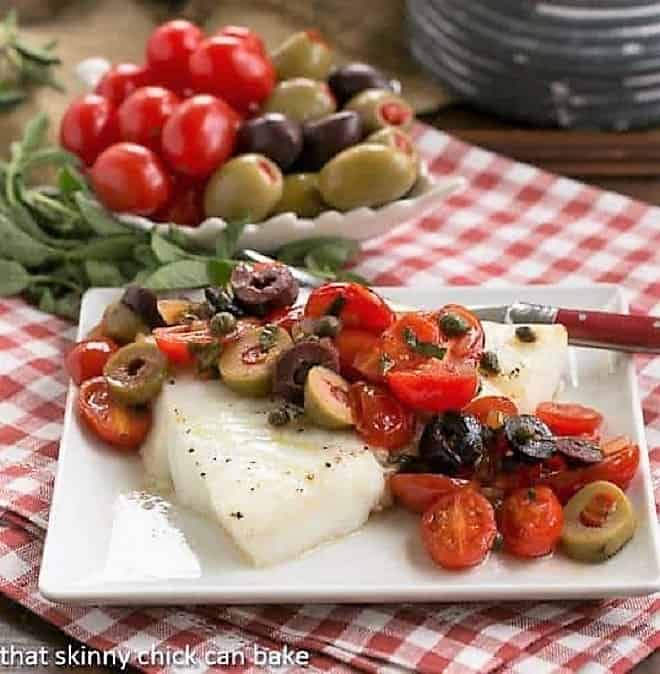 Sea Bass with Tomatoes, Olives and Capers - A simple roasted fish recipe with a quick Mediterranean topping. Sea Bass with Tomatoes, Olives and Capers on small square plate with fork.