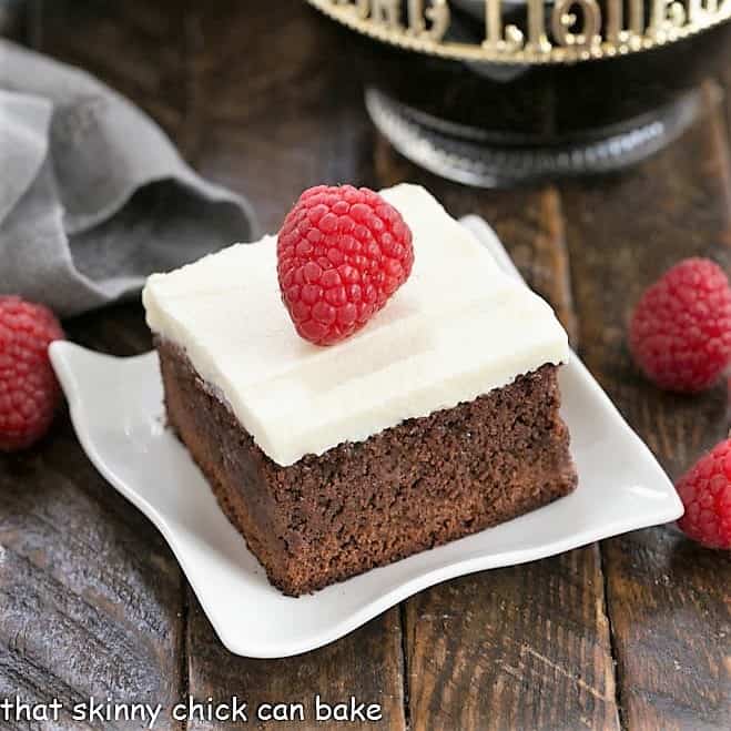 A raspberry brownie topped with cream cheese frosting and a fresh raspberry on a white plate.