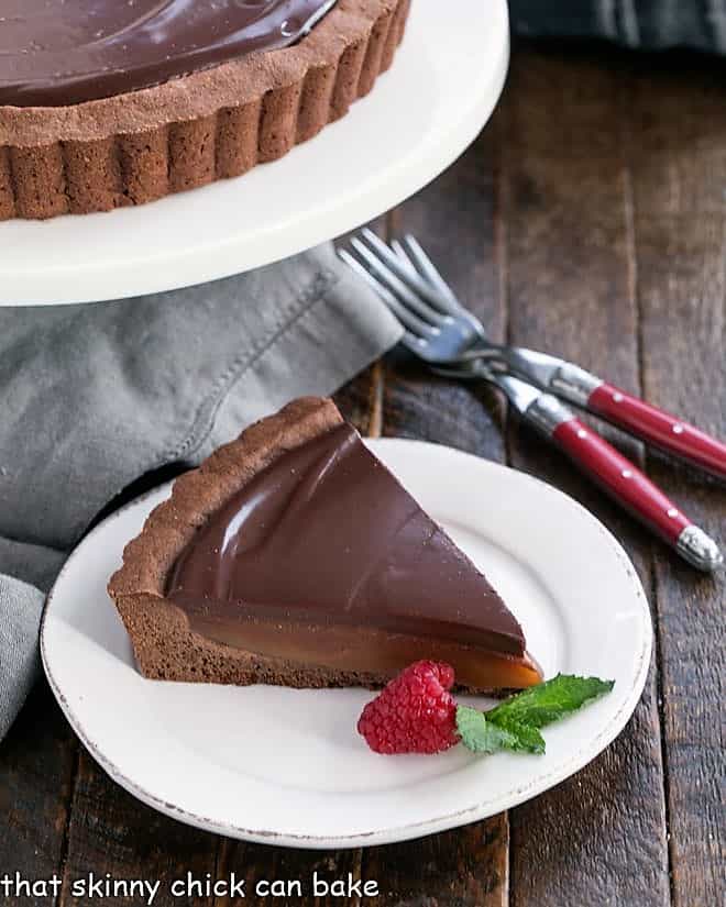 Chocolate Caramel Tart slice on a white plate next to a cake stand and two forks.