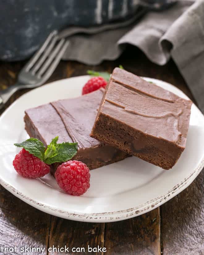 2 poke brownies on a white dessert plate with raspberries and mint to garnish.