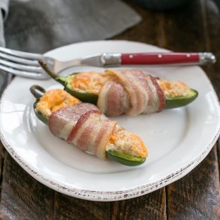 2 bacon wrapped jalapeno poppers on a white ceramic plate with a red handled fork