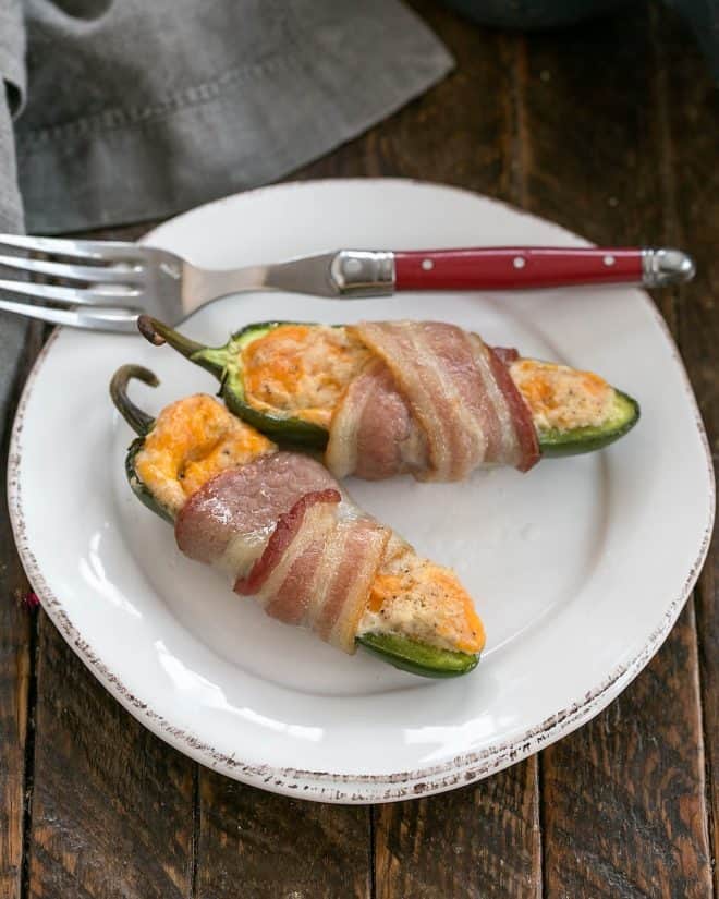Two bacon wrapped jalapeno poppers on a round white plate with a red handle fork.