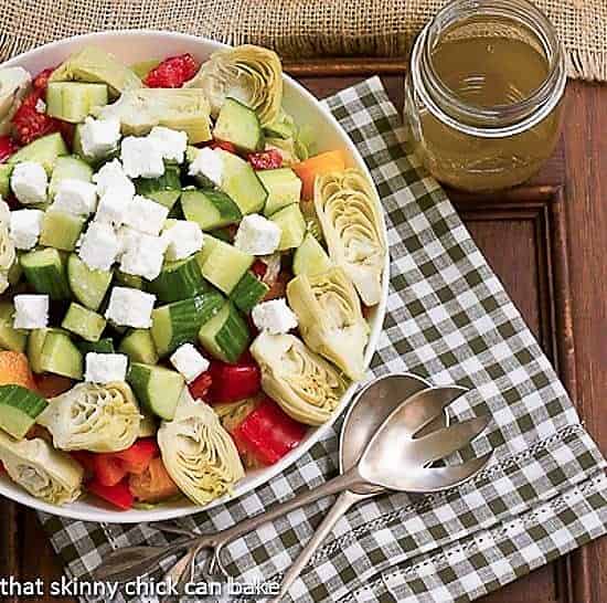 Greek Salad with Feta in a serving bowl with salad serving utensils and a jar of vinaigrette.