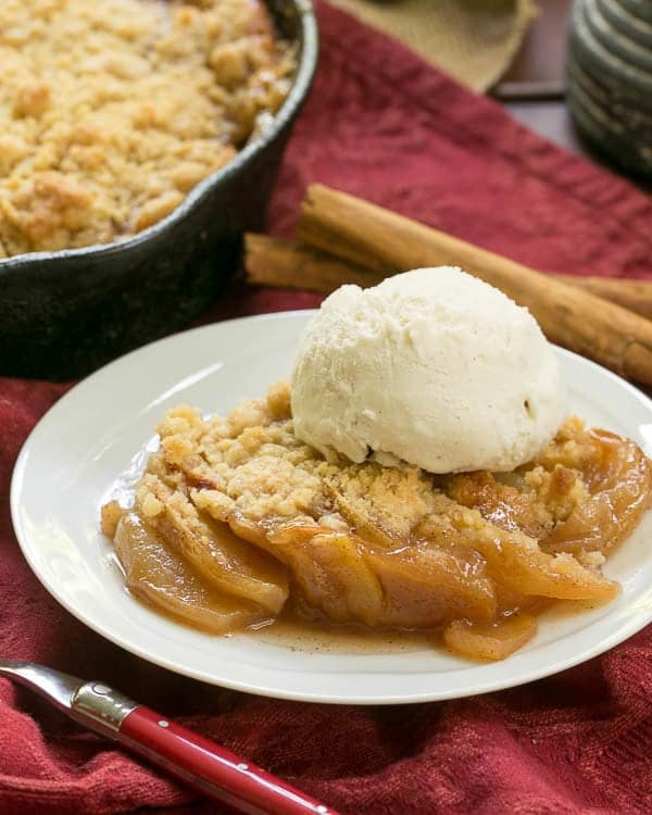 Grilled Apple Crisp on a white dessert plate topped with a scoop of ice cream and in front of a cast-iron skillet of apple crisp.