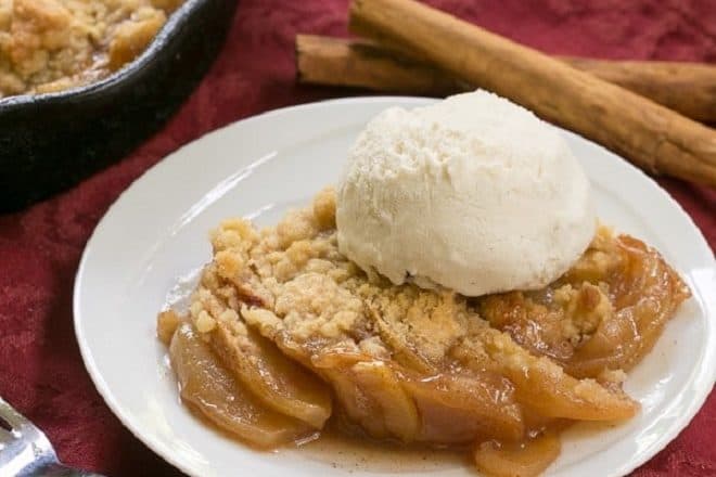 Grilled Apple Crisp topped with vanilla ice cream with a red fork and cinnamon sticks
