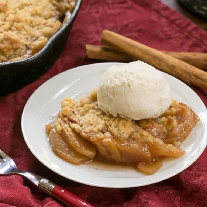 Grilled Apple Crisp topped with vanilla ice cream with a red fork and cinnamon sticks.