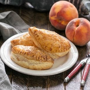 3 peach hand pies on a round white plate with 2 peaches in the background