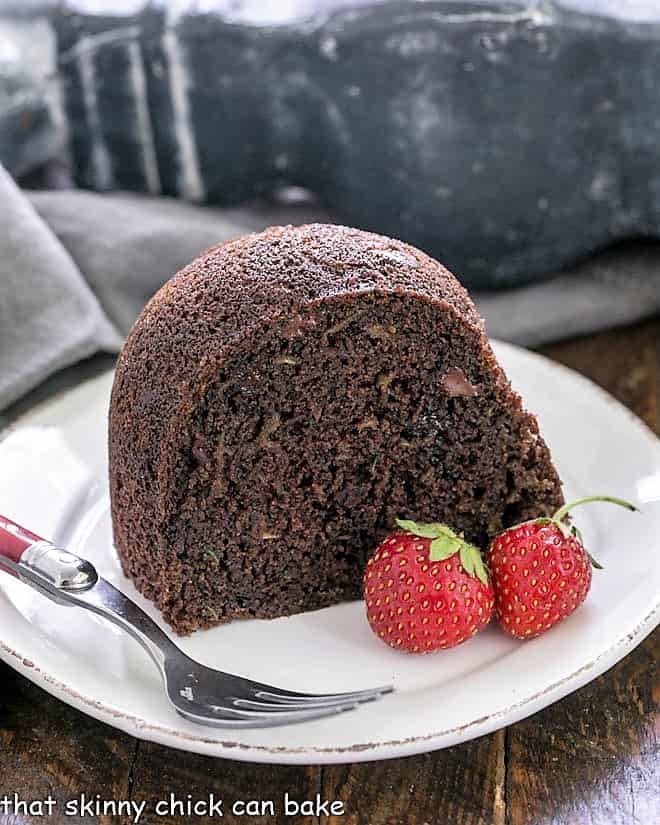 Slice of Chocolate Zucchini Bundt Cake on a white plate with a red handle fork and two strawberries.