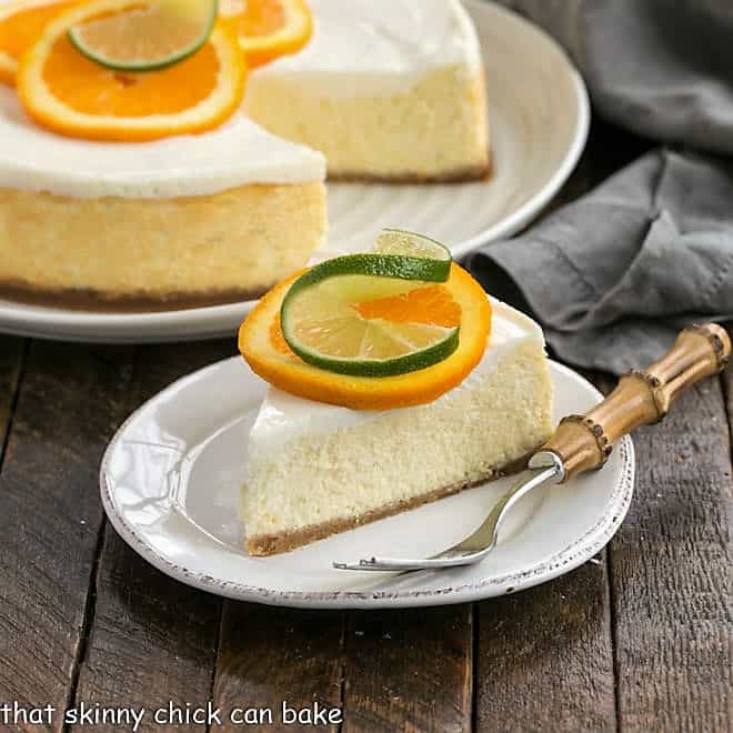  Margarita Cheesecake slice on a white dessert plate garnished with an orange and lime slice in front of the dessert with a slice remove