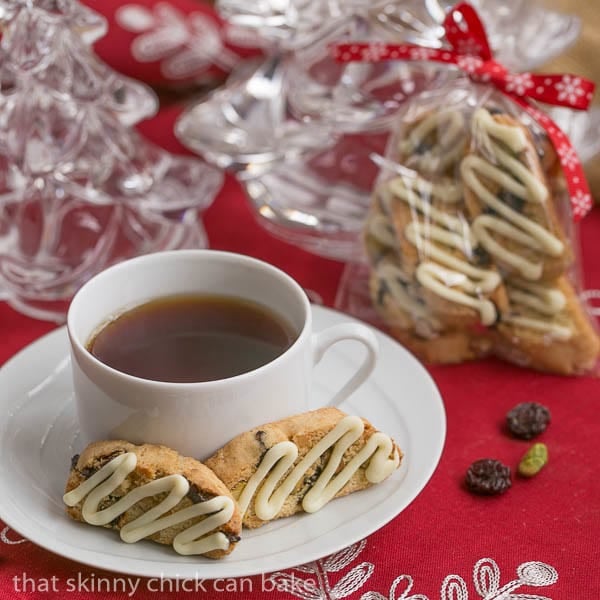 Cherry Pistachio Biscotti sits on a white saucer with a cup of tea. Bags of biscotti are festively wrapped for gifts in the background
