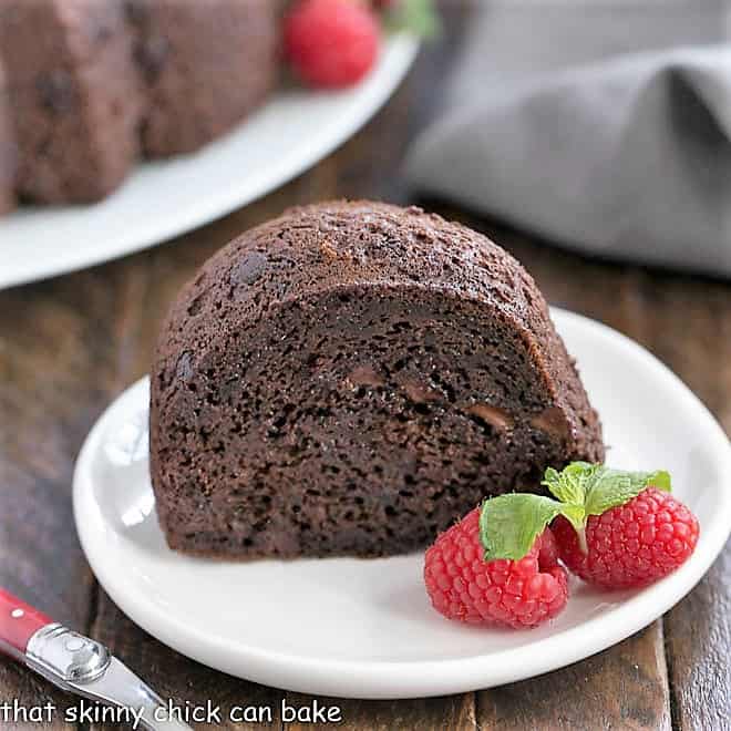 This Easy Chocolate Kahlua Cake is rich, dense and irresistible! Made with a cake mix, no one will ever guess it's not made from scratch until you share the recipe! Slice of Kahlua Bundt Cake on a white dessert plate with two raspberries and a sprig of mint.