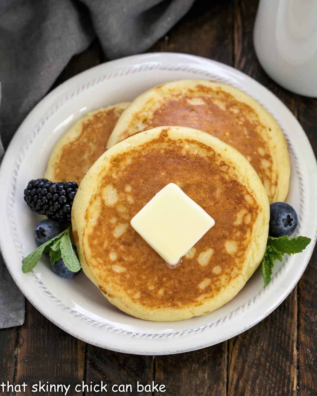 Overhead view of 3 buttermilk pancakes topped with butter and garnished with berries and mint.