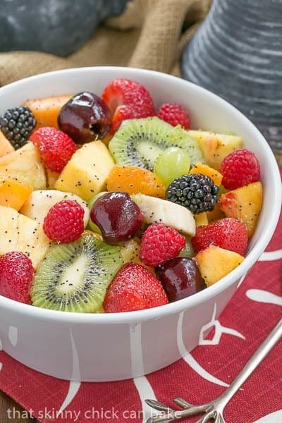 A white bowl of colorful fresh fruit with poppyseet dressing on a red and white napkin.