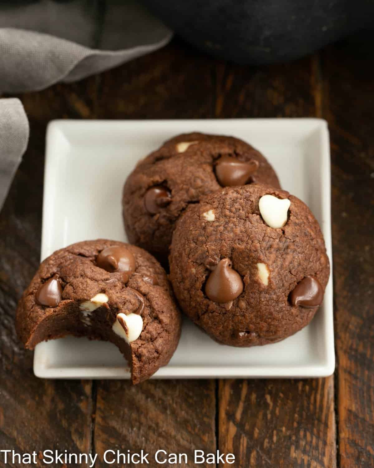 3 chocolate chocolate chip cookies on a square white plate, one with a bite removed.