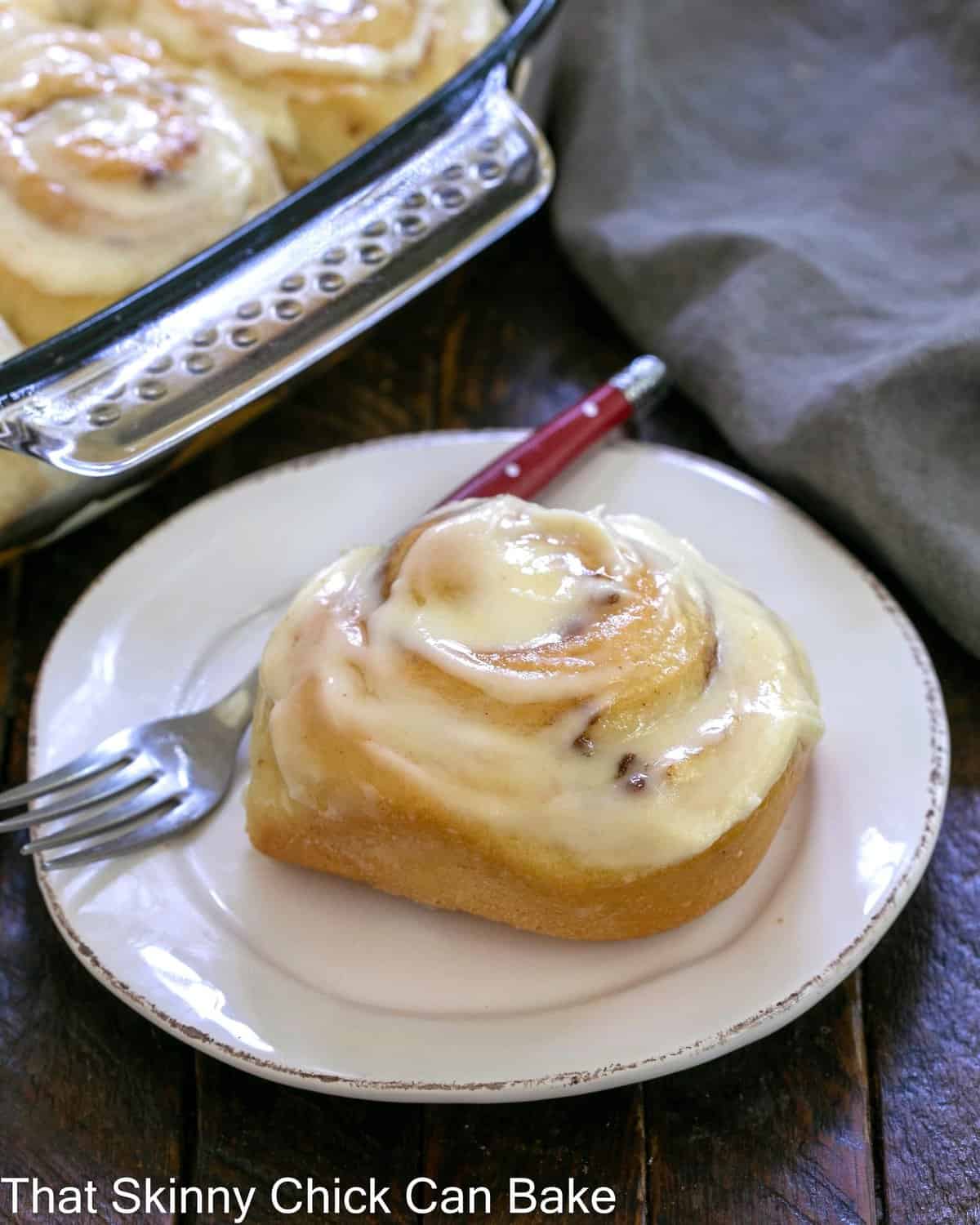 One cinnamon roll on a white plate in front of pan of rolls.