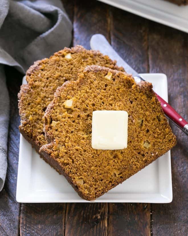 2 slices of pumpkin bread with apples on a square white plate with a butter knife.
