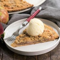 Apple Pie Pizza slice with a scoop of ice cream on a white dessert plate with a red handled fork in front of whole pizza