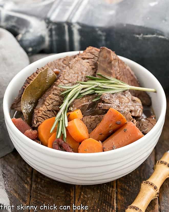 Beef brisket recipe in a white ceramic bowl with carrots and rosemary.