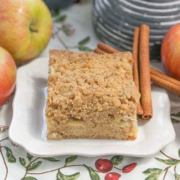 Apple Coffee Cake slice on a white plate surrounded by fresh apples and cinnamon sticks