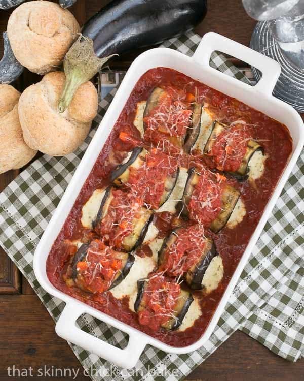 Eggplant Rollatini in a white casserole dish surrounded by wheat rolls and a whole eggplant 