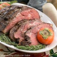 Classic Prime Rib slices next to the roast on a white platter with rosemary and persimmon garnishes