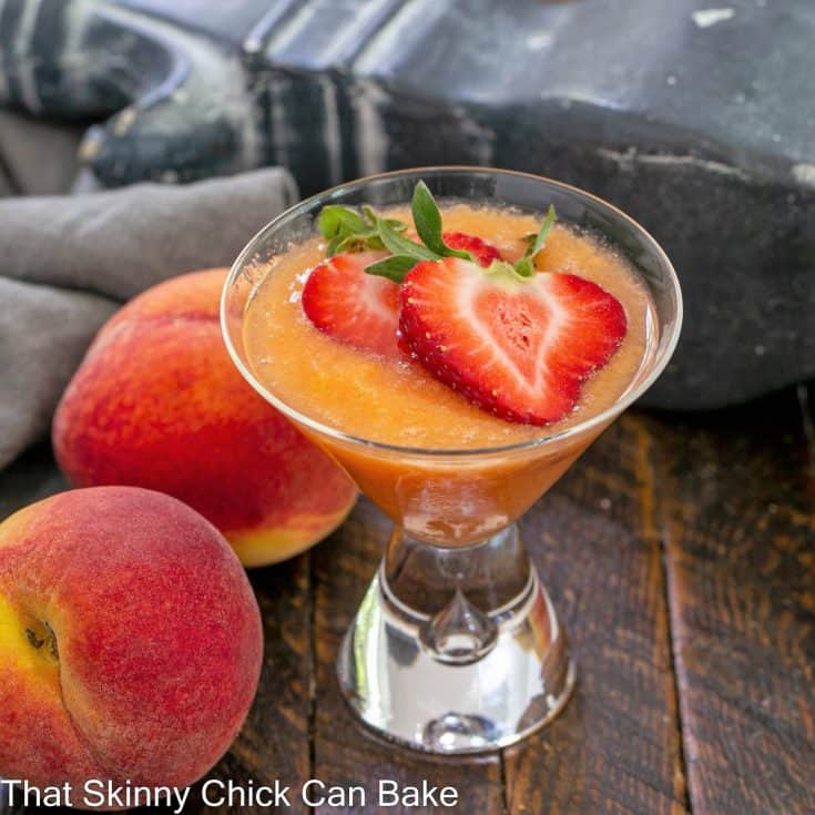 A peach bellini recipe topped with sliced strawberries next to 2 fresh peaches