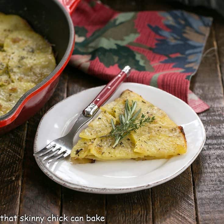 Slice of potato galette garnished with herbs on a round white plate with a red handle fork