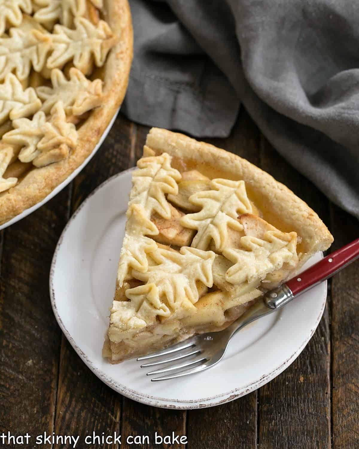 Overhead of a slice of apple pie on a round white dessert plate next to a partial view of the rest of the pie.