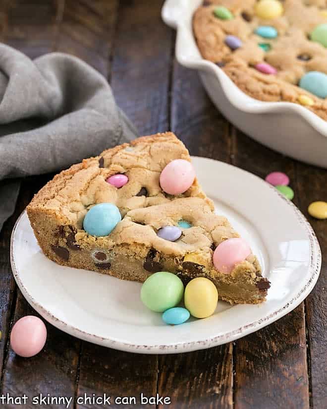 Slice of cookie cake on a white plate with the rest of the cookie cake in the background.