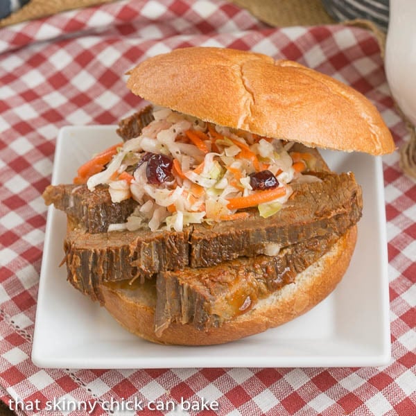 Oven Braised Texas Brisket sandwich topped with coleslaw sits on a white plate with a red and white gingham tablecloth underneath