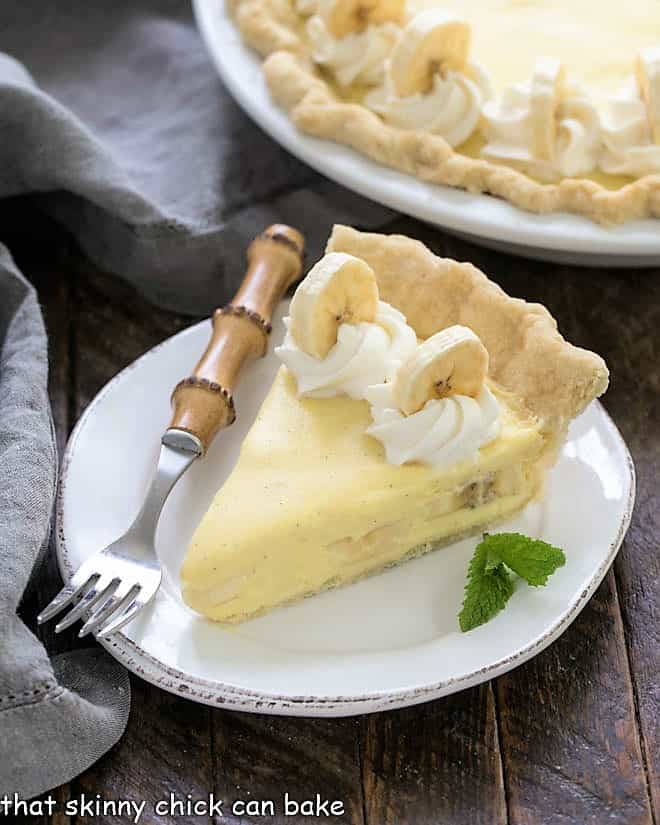 Slice of banana cream pie on a white plate with a bamboo handle fork with a glimpse of the whole pie in the background.