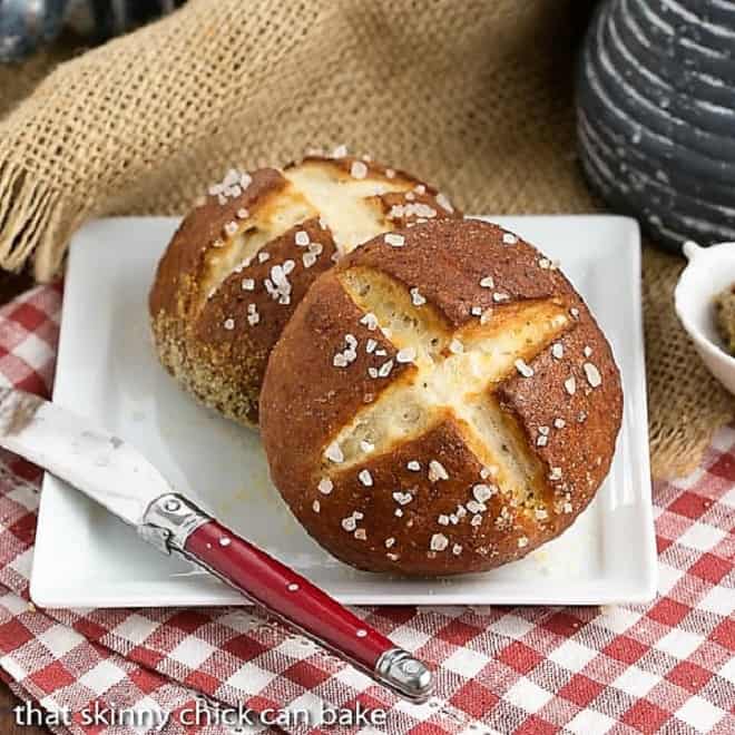 Two Pretzel Rolls on a square white plate with a red handled knife.