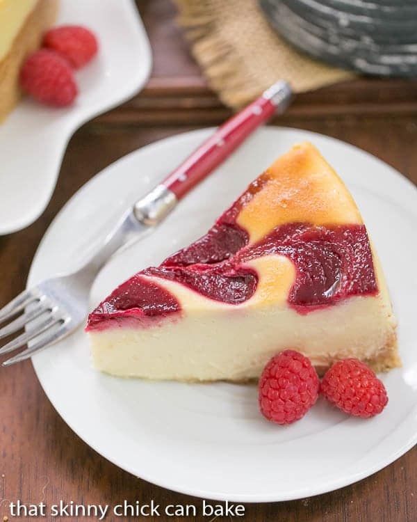 White Chocolate Raspberry Swirl Cheesecake - A luscious White Chocolate Cheesecake with a marbleized raspberry topping White Chocolate Raspberry Swirl Cheesecake slice on a white plate with a red handled fork.