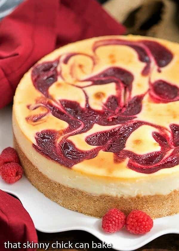 White Chocolate Raspberry Swirl Cheesecake - A luscious White Chocolate Cheesecake with a marbleized raspberry topping White Chocolate Raspberry Swirl Cheesecake on a white plate garnished with fresh raspberries.