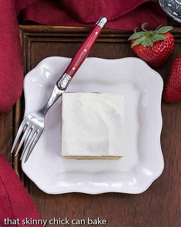 Overhead view of a slice of white sheet cake on a white plate with a red handled fork