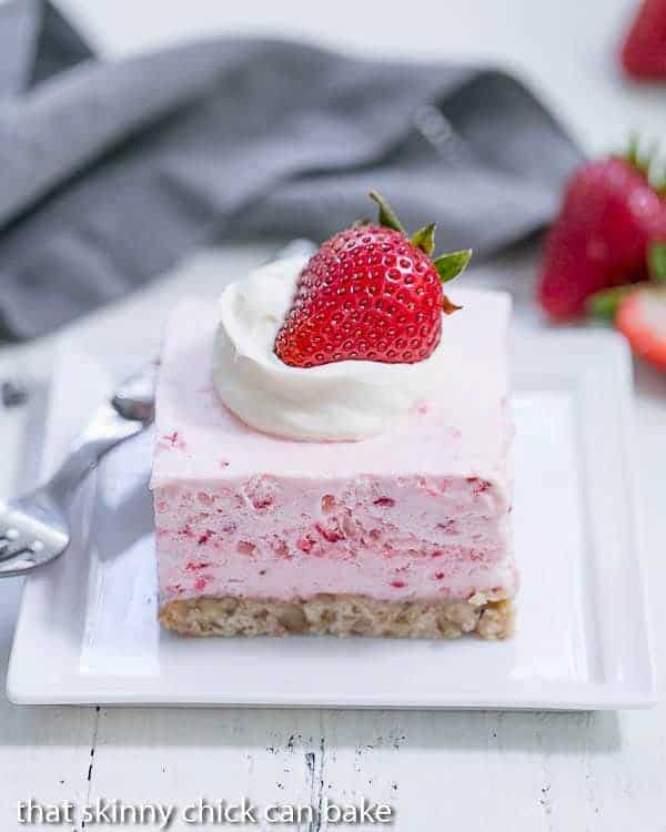 En skive Strawberry Pie Dessert på en firkantet hvid tallerken toppet med fløde og et halvt jordbær.