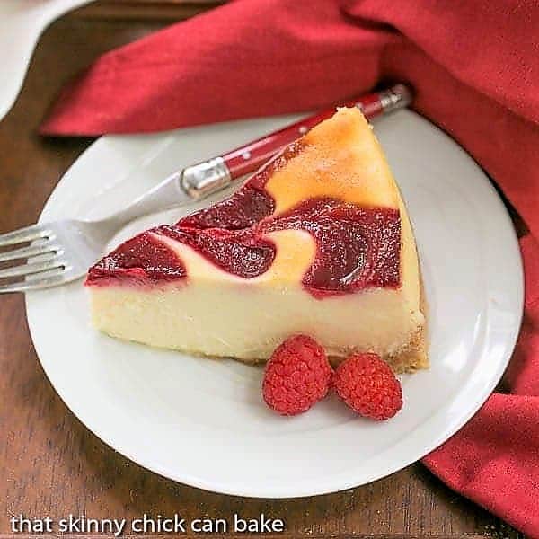 White Chocolate Raspberry Swirl Cheesecake slice on a round white plate with fresh raspberries and a red handle fork