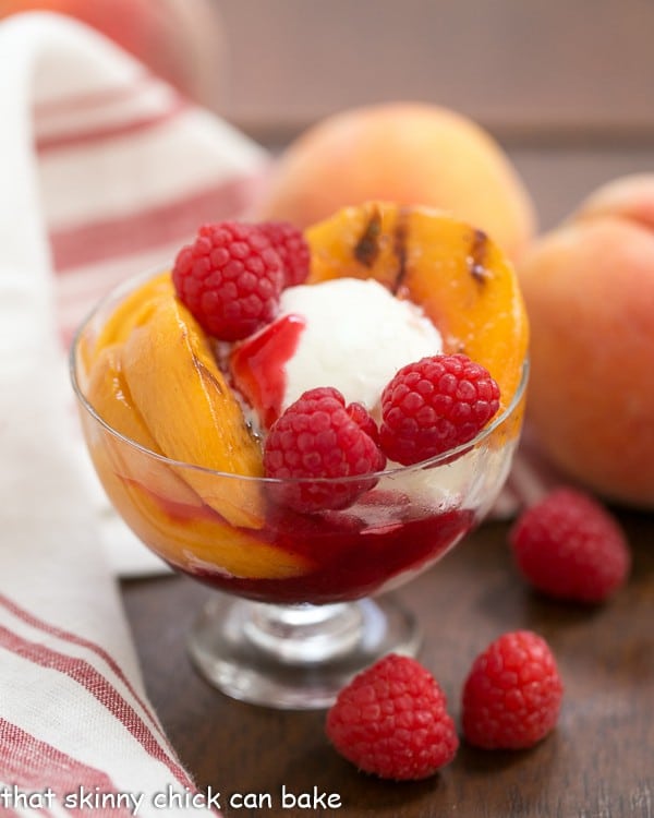 Grilled Peach Melba in a glass serving dish with a red white napkin, peaches, and fresh raspberries.