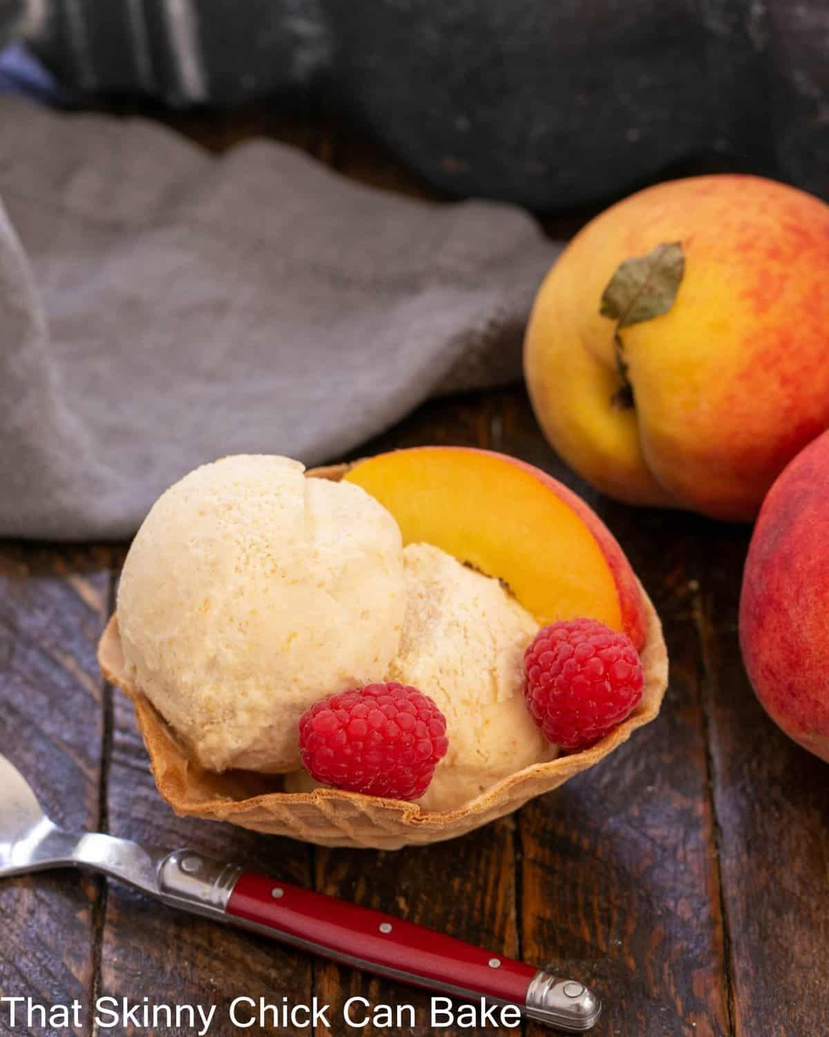 No-churn peach ice cream in a sugar cone bowl with raspberry and peach garnishes.