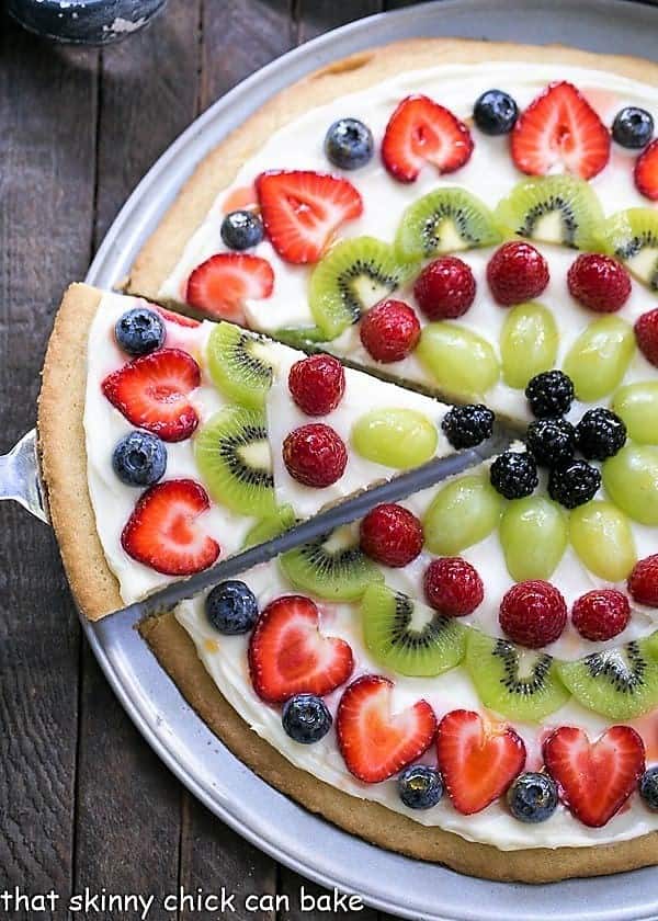 Overhead view of fresh fruit pizza with a slice being removed.