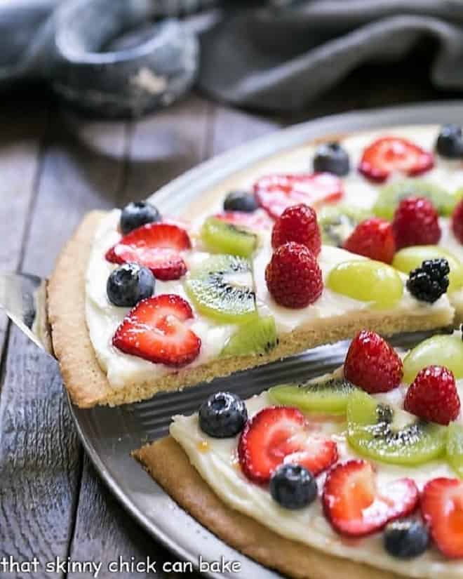 A slice of Fresh Fruit Pizza being removed from a whole pizza on a metal pizza pan.