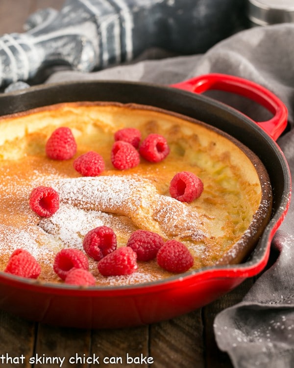 Best Dutch Baby Pancake in a red cast iron skillet topped with fresh raspberries and a dusting of powdered sugar.