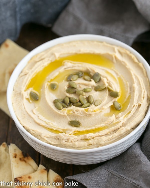 White bowl of creamy homemade hummus garnished with olive oil and pepitas.