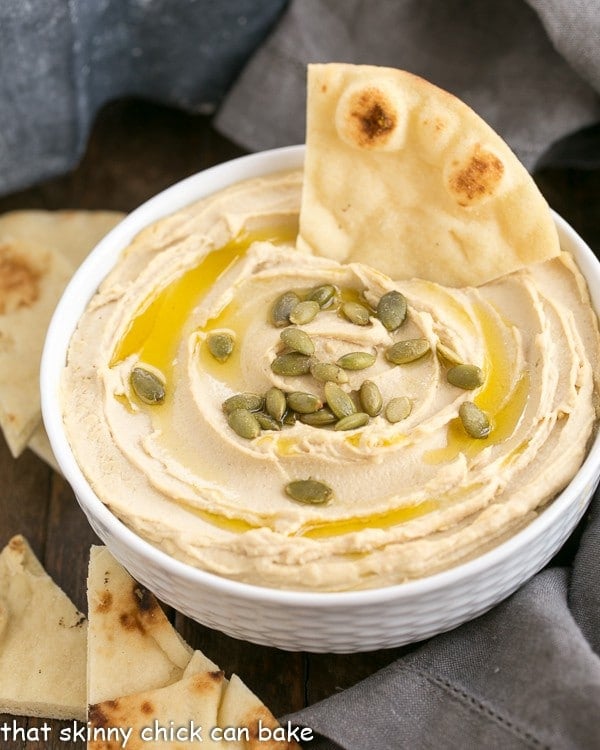 A white bowl of creamy hummus from scratch, garnished with pepitas and a drizzle of olive oil.