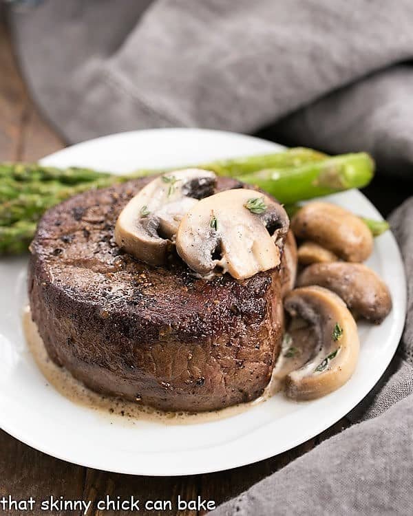 Mushroom Topped Pepper Steak topped with mushrooms on a white plate with asparagus spears