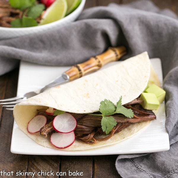 Slow Cooker Carne Asada in a flour tortilla on a square white plate with a bamboo handle fork.